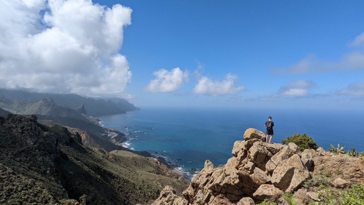 Looking out across cliffs, out toward the ocean. The sun is shining, the sky is bright blue, with just a few clouds in the sky